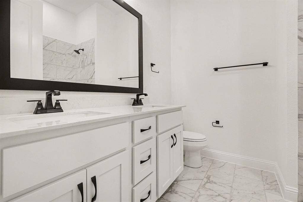 Double vanity with white cabinetry, stone-finish countertops, matte black fixtures, framed mirror, and stone-tile flooring