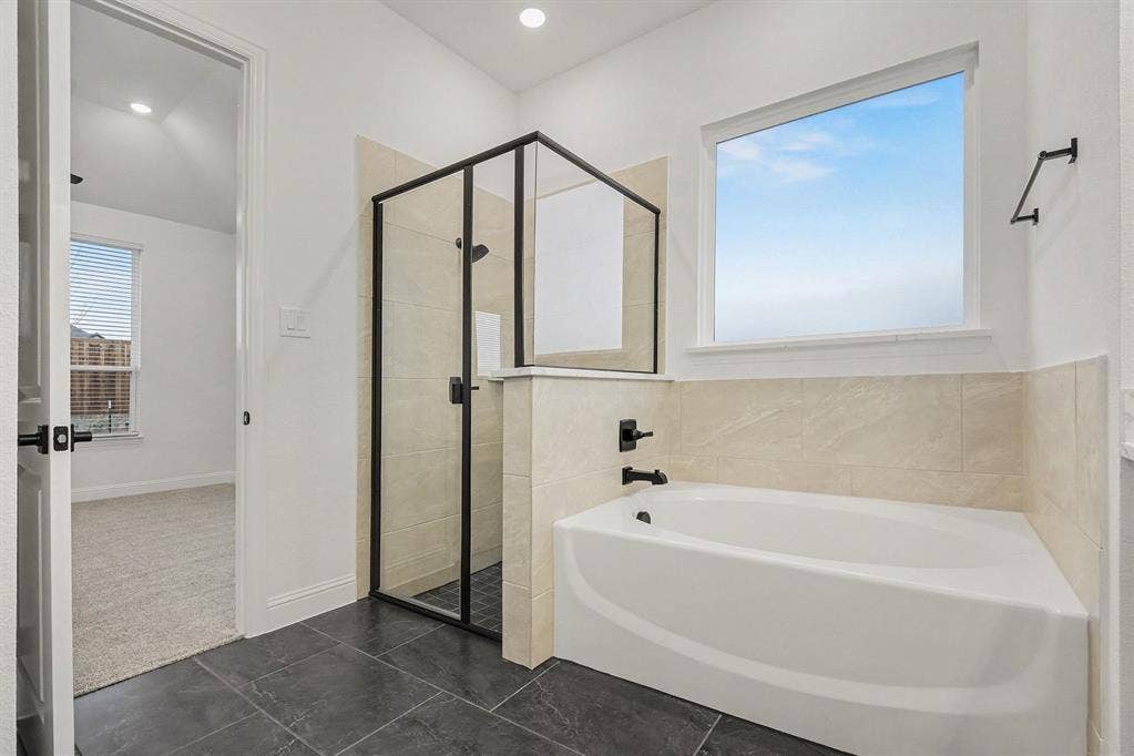 Soaking tub with a wall-mounted faucet, separate shower enclosure with a matte black frame, neutral-toned tiled surround, and dark floor tiling