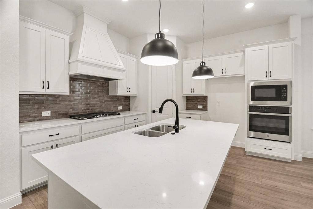 Kitchen featuring a large central island with light-toned solid surface countertops, custom white cabinetry, built-in stainless steel double ovens, tile backsplash, and matte black pendant lighting