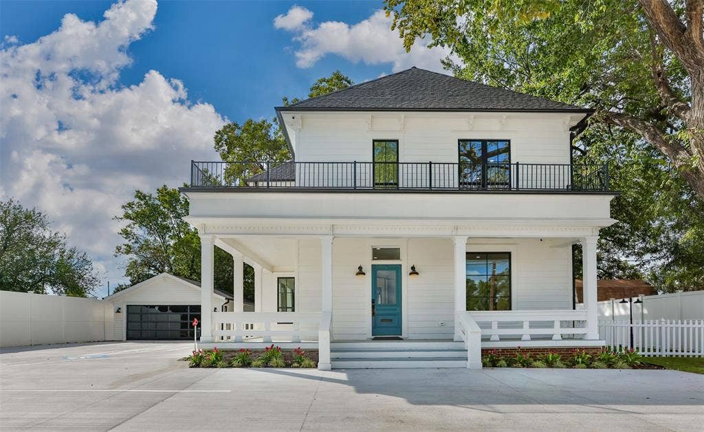 Two-story exterior featuring white horizontal siding, a covered front porch with decorative millwork, a second-story balcony with black metal railing, and a detached garage