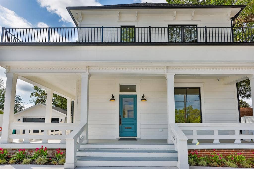 Two-story exterior featuring white horizontal siding, a covered front porch with a wood-style railing, a second-story balcony with metal railings, and a blue painted front door