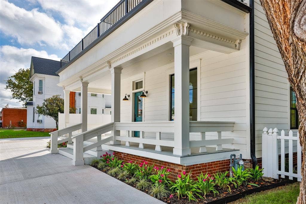 Covered front porch featuring white horizontal siding, decorative millwork columns, a brick foundation, and an upper-level balcony with metal railing