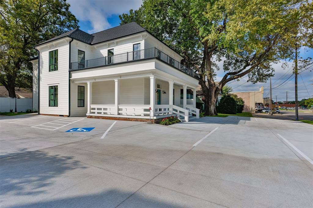 Two-story exterior featuring white horizontal siding, a wraparound covered porch, second-story balcony with metal railing, and concrete parking area