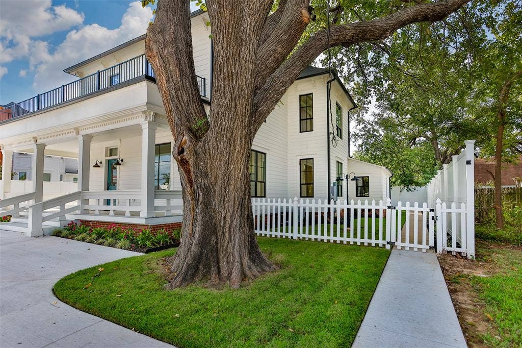 Two-story exterior featuring white horizontal siding, a covered front porch with a brick base, second-level balcony with black railing, and white picket fencing
