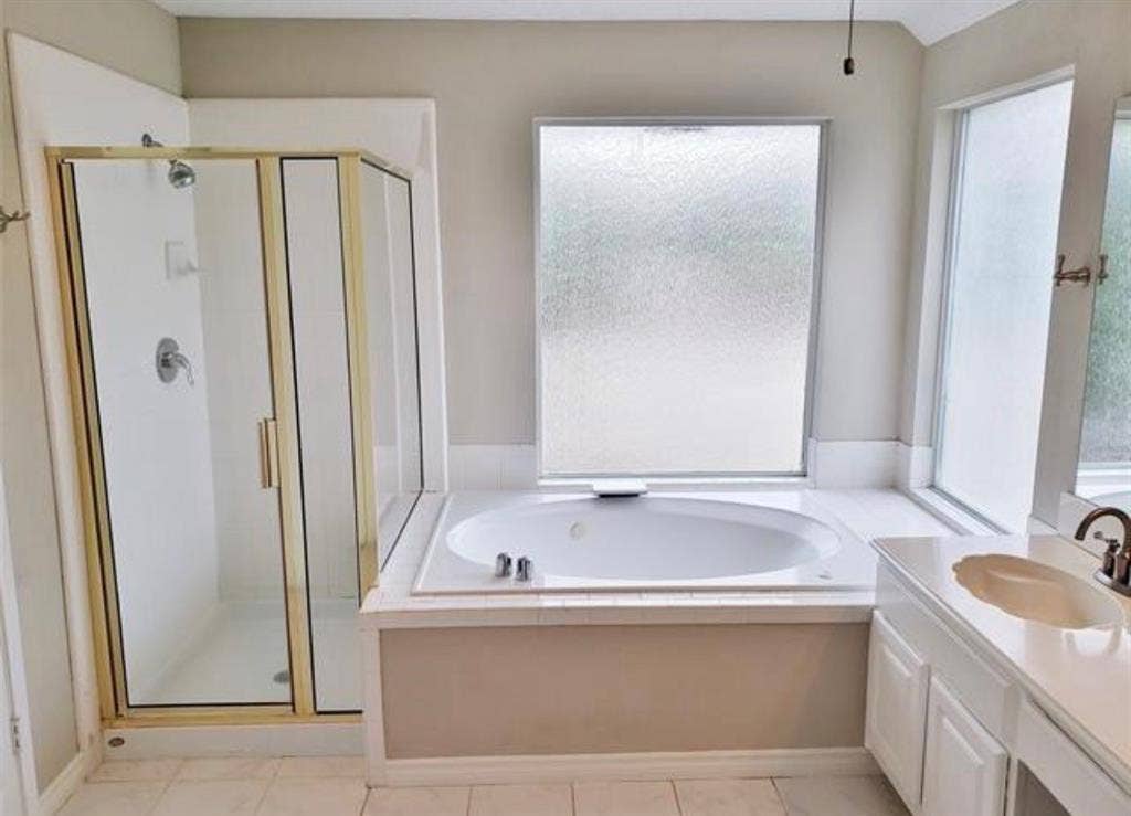 211 Moss Hill Road, Irving, TX 75063 White tile flooring with an embedded tub, an enclosed shower with gold-tone framing, and a vanity with an integrated sink