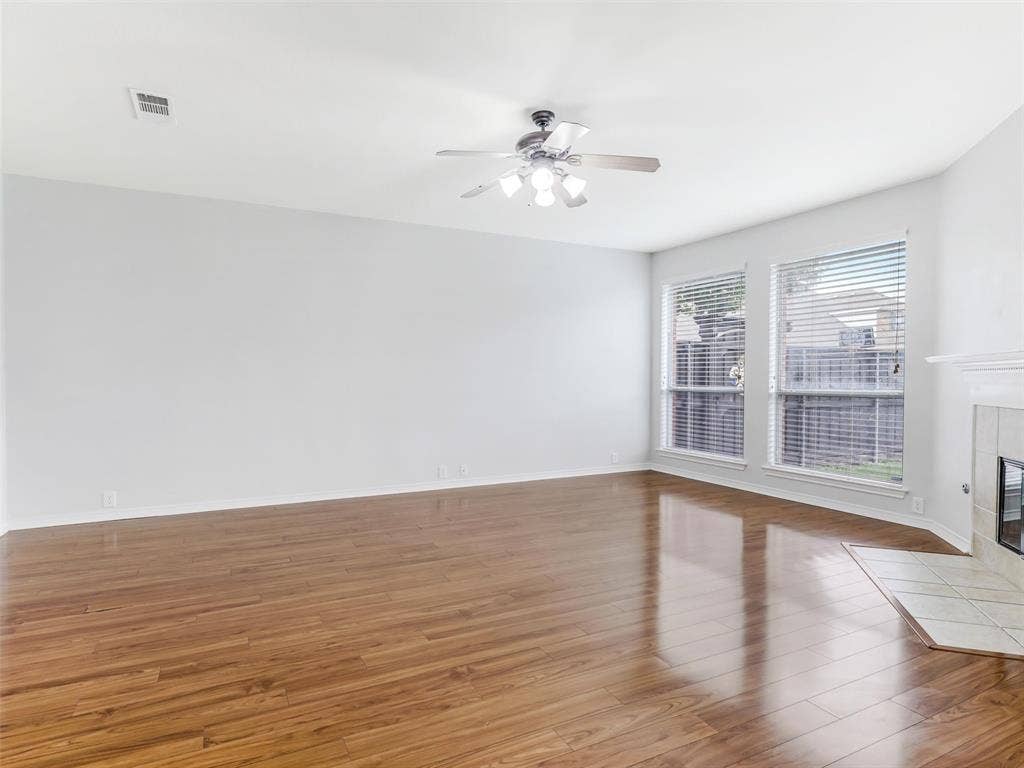 7419 Ravenhill Drive, Frisco, TX 75035 Wood-finish flooring, white walls, ceiling fan with integrated lighting, two windows with blinds, and a tiled fireplace hearth