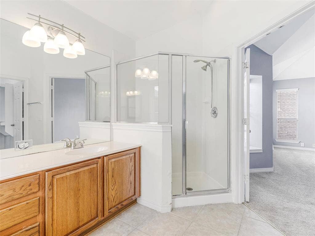 7419 Ravenhill Drive, Frisco, TX 75035 Light-toned tile flooring, wood-finish vanity with an integrated sink, full-width mirror, multi-bulb vanity light fixture, and an enclosed shower