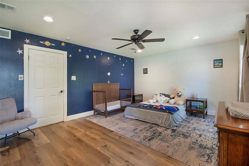 4074 Cedar Bayou Drive, Dallas, TX 75244 Spacious bedroom featuring warm wood-finish flooring, a ceiling fan with dark metal blades, recessed lighting, and a deep navy accent wall adorned with decorative celestial details