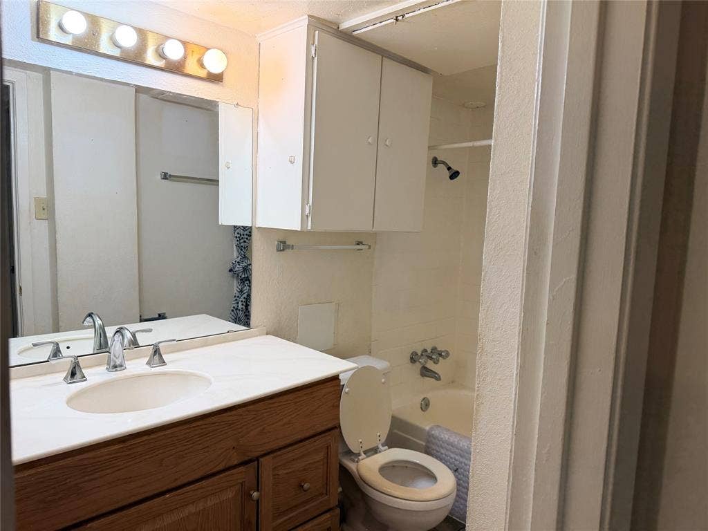 12888 Montfort Drive, #167, Dallas, TX 75230 Bathroom featuring a wood-finish vanity with an integrated sink and chrome faucet, a large mirrored wall, overhead cabinetry, and a bathtub with an integrated shower head