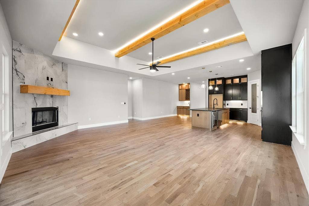 Light wood-finish flooring, vaulted ceiling with exposed wood beams and recessed lighting, tiled fireplace with wood mantel, kitchen island with built-in beverage refrigerator, and dark wood cabinetry