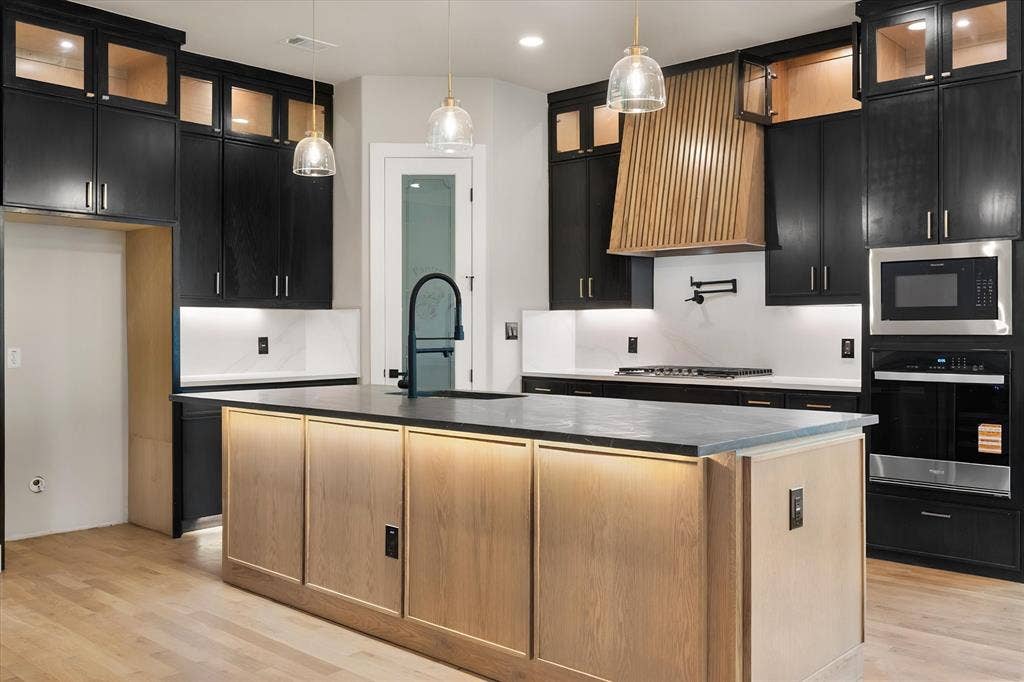 Kitchen featuring a large island with integrated lighting, a custom wood hood, stainless steel appliances, black cabinetry, and light wood-finish flooring