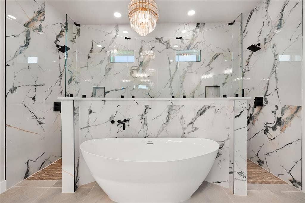 Freestanding soaking tub with a wall-mounted faucet, dual walk-in showers with glass enclosures, full-height tile surround, and a crystal chandelier