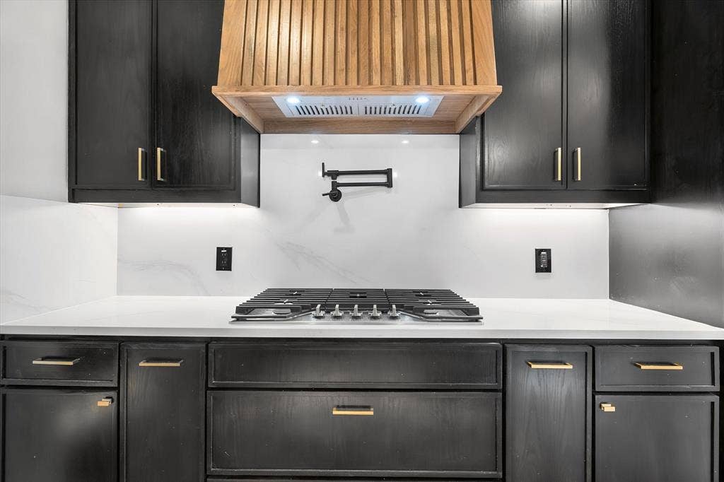 Custom wood range hood, black cabinetry with gold-tone hardware, white countertops, coordinating white backsplash, and gas cooktop