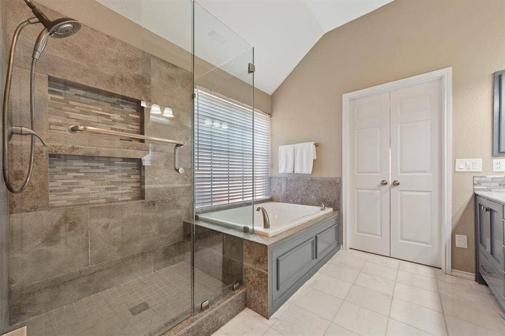 Walk-in shower with glass enclosure and mosaic tile accents, soaking tub with tiled surround, dual panel bifold doors, and a vaulted ceiling