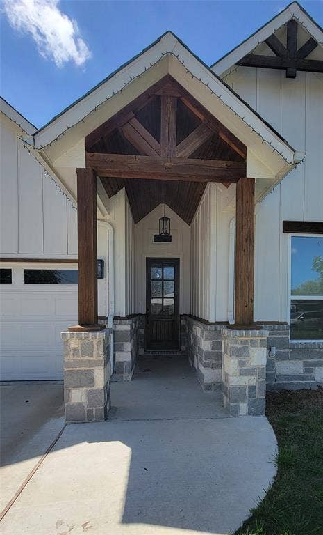 3207 NW 30th Street, Fort Worth, TX 76106 Covered entryway with robust wood beams, stone pillar bases, vertical plank siding, a glass-paned entry door, and an attached outdoor light fixture