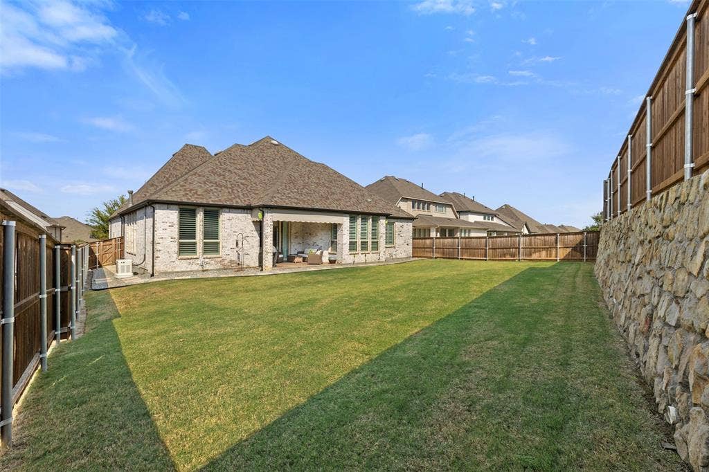 9716 Wilderness Road, Little Elm, TX 75068 Expansive backyard featuring a manicured lawn, a stone retaining wall, and a covered patio with recessed lighting