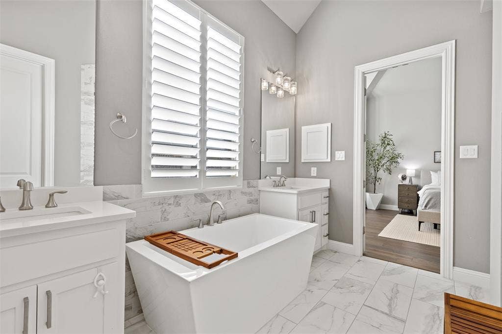 9716 Wilderness Road, Little Elm, TX 75068 Freestanding soaking tub, white plantation shutters, dual vanities with white cabinetry, wall-mounted mirror, and tile flooring