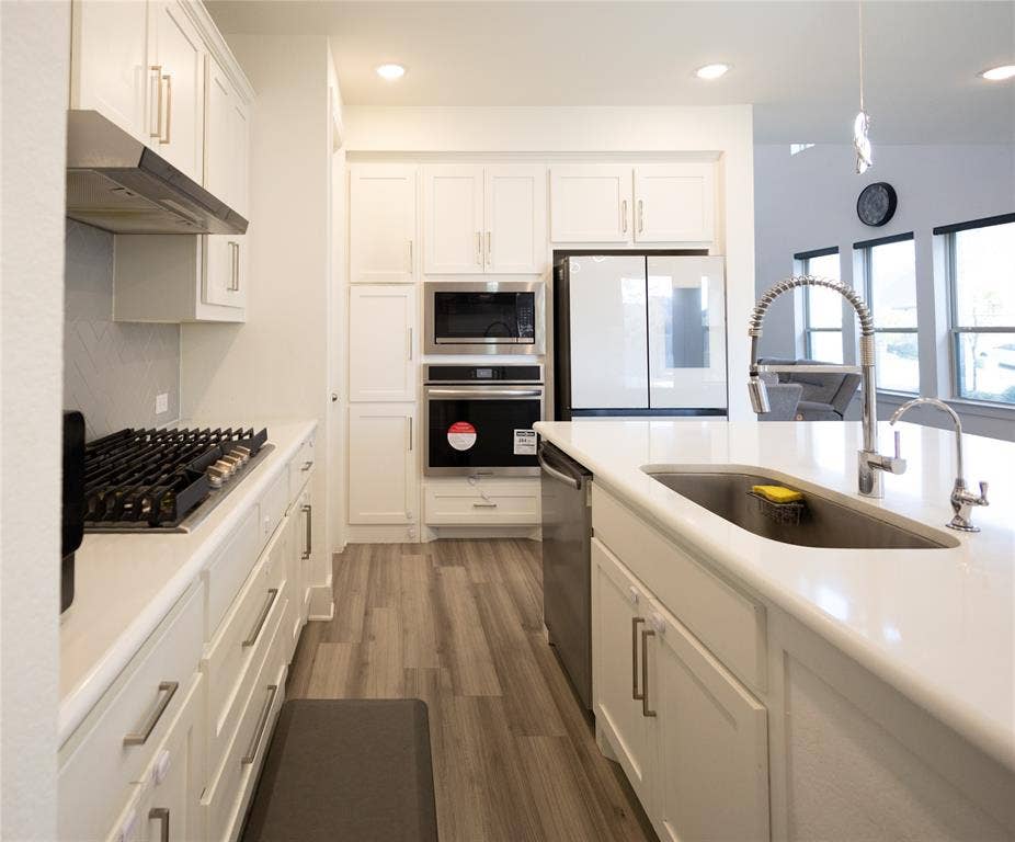 3017 Andorra Road, McKinney, TX 75071 Modern galley kitchen featuring white cabinetry with chrome hardware, wood-finish flooring, quartz countertops, dual stainless steel ovens, gas cooktop, stainless steel refrigerator with ice dispenser, and an oversized apron-front sink with contemporary arc faucet