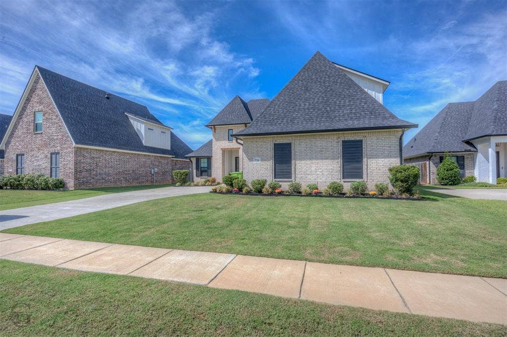 Cream brick exterior with dark shingle roofing, prominent gables, a concrete driveway, and a manicured lawn