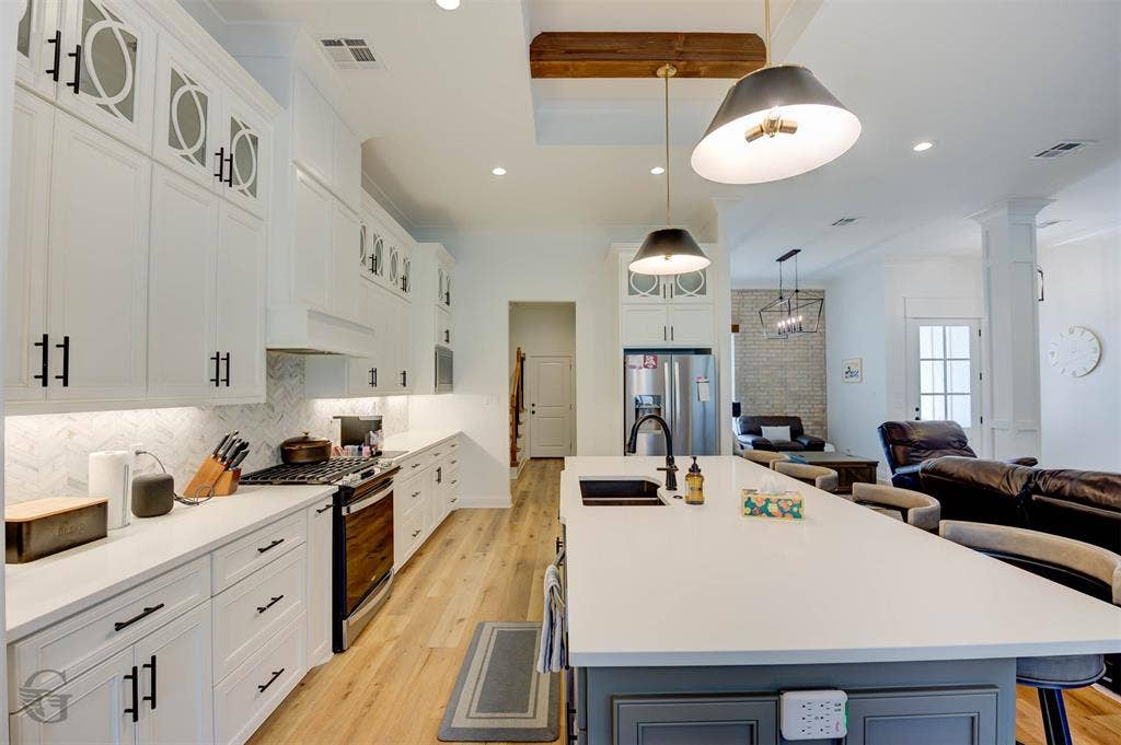 White cabinetry with decorative glass inserts, stainless steel appliances, a large center island with a built-in sink, wood-finish flooring, and a recessed lighting fixture