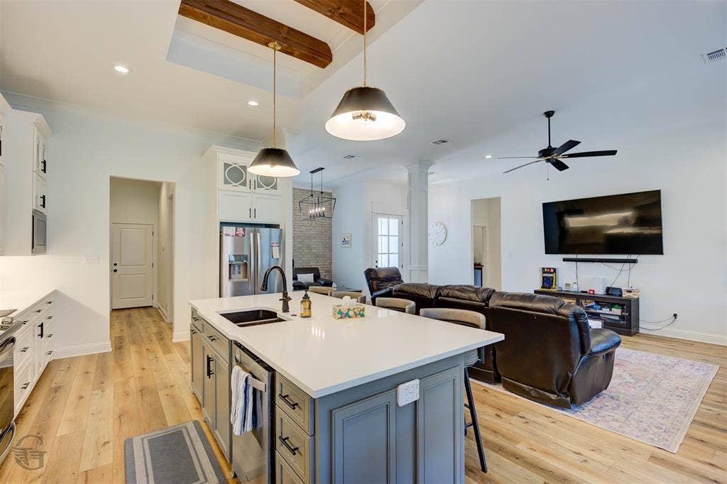 Open-concept kitchen and living area featuring a blue-gray island with white quartz countertop and pendant lighting, stainless steel refrigerator, wood-finish flooring throughout, exposed beam ceiling details, ceiling fan, wall-mounted television, and abundant natural light from multiple windows