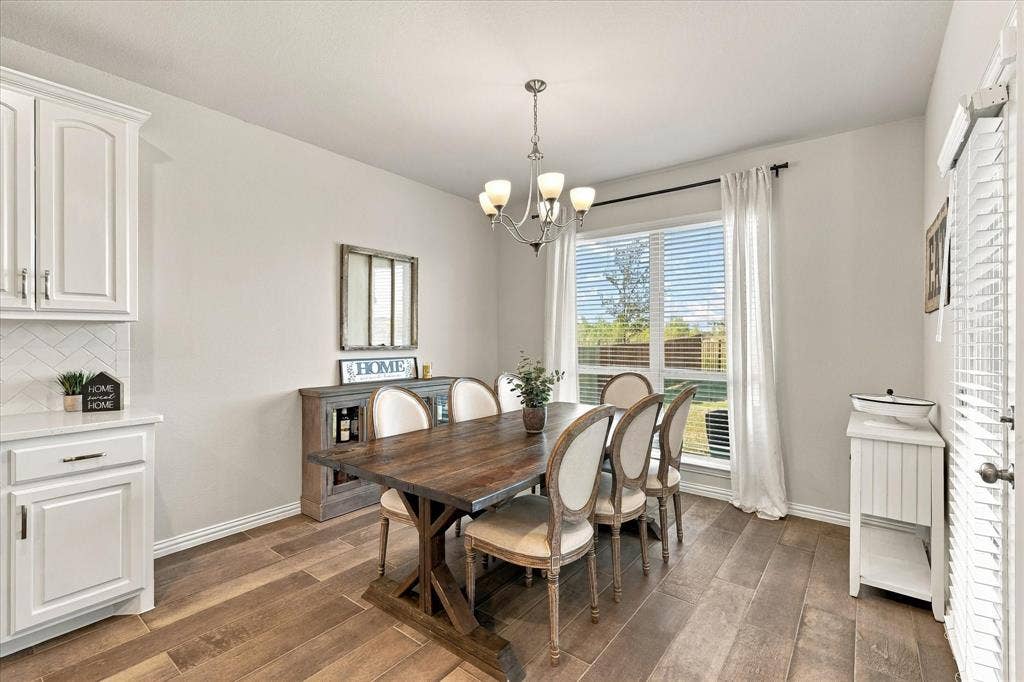 Wood-finish flooring, white cabinetry with hardware, herringbone tile backsplash, large window with blinds, and a chandelier lighting fixture