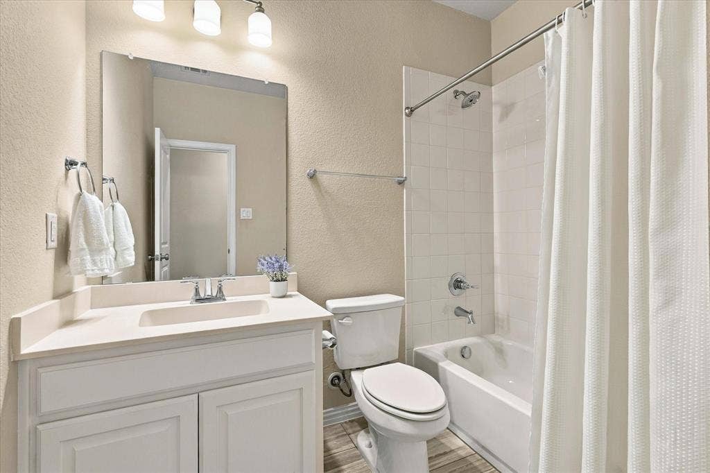 White vanity with integrated sink, rectangular mirror, three-light vanity fixture, white tiled tub/shower combination, and wood-finish flooring