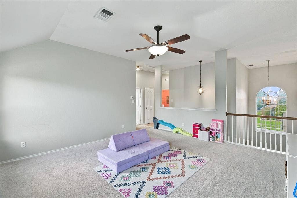 Vaulted ceilings, neutral carpeting, a central ceiling fan with integrated lighting, a half-wall partition with pendant lighting, and an arched window with a wooden railing