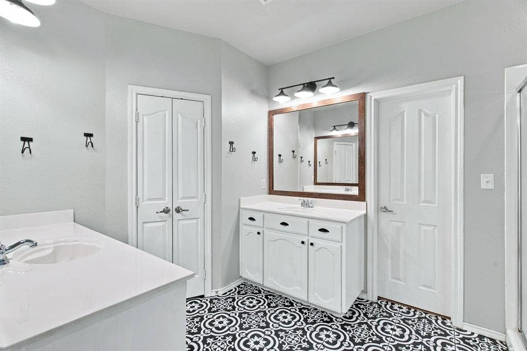 Bathroom featuring dual white vanities with solid-surface countertops, pattern-tiled flooring, three-light wall-mounted fixtures, and wood-framed mirrors