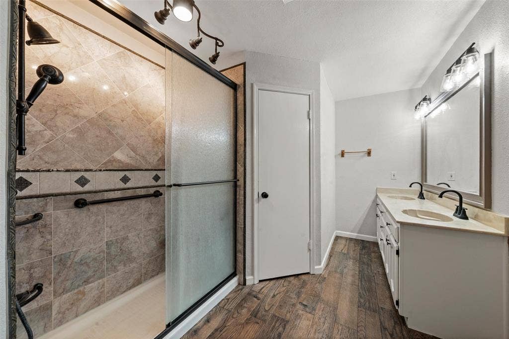 812 Regina Drive, Hewitt, TX 76643 Walk-in shower with tile surround and dark bronze fixtures, dual vanity with undermount sinks and light-toned countertops, wood-finish flooring, and track lighting