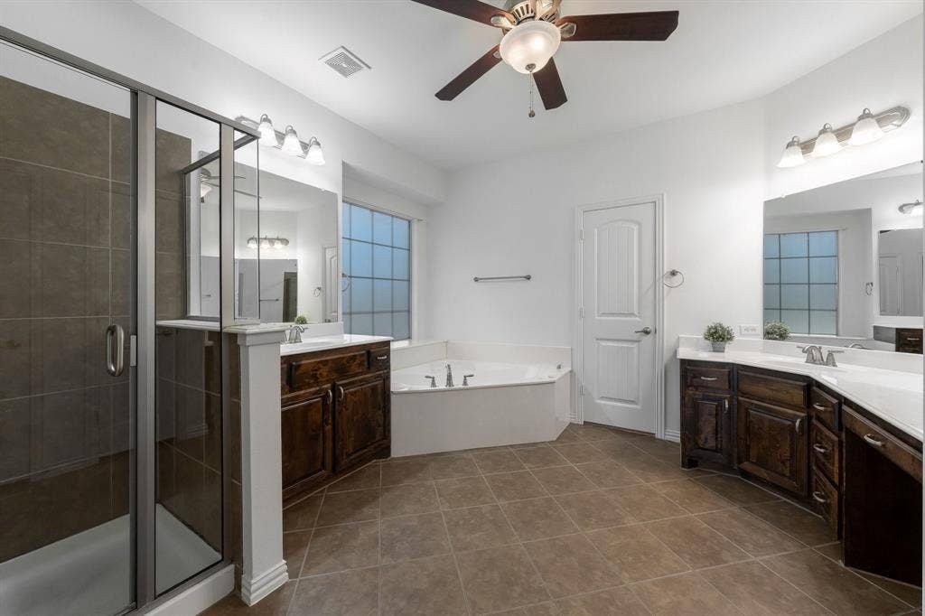 Spacious bathroom featuring a glass-enclosed shower, corner soaking tub, dual vanities with dark wood cabinetry, and a ceiling fan