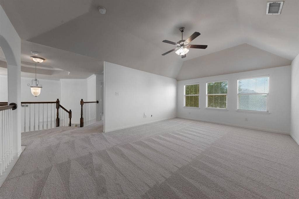 Vaulted ceiling with a ceiling fan, three windows with blinds, light-toned carpeting, and a staircase with white spindles and dark wood banisters