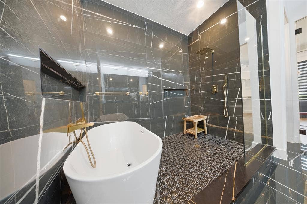 522 S Clark Road, Cedar Hill, TX 75104 Freestanding soaking tub with a gold-tone floor-mounted faucet, walk-in shower with a glass enclosure, black veined wall tiles, recessed shelving, and a patterned shower floor