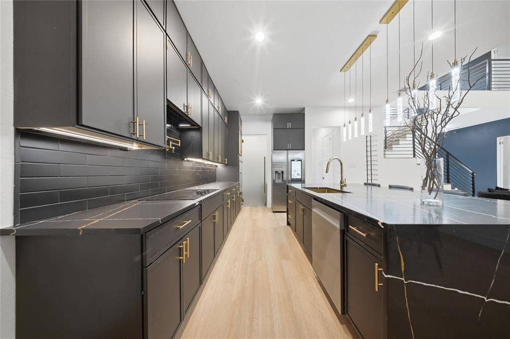 522 S Clark Road, Cedar Hill, TX 75104 Modern kitchen featuring dark cabinetry with gold-tone hardware, black subway tile backsplash, light wood-finish flooring, and a large island with a gold-veined countertop and integrated sink