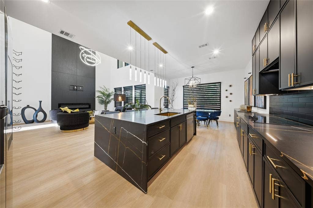 522 S Clark Road, Cedar Hill, TX 75104 Light wood-finish flooring, dark kitchen cabinetry with gold-tone hardware, a large kitchen island with a stone countertop, a linear pendant light fixture, and a built-in fireplace