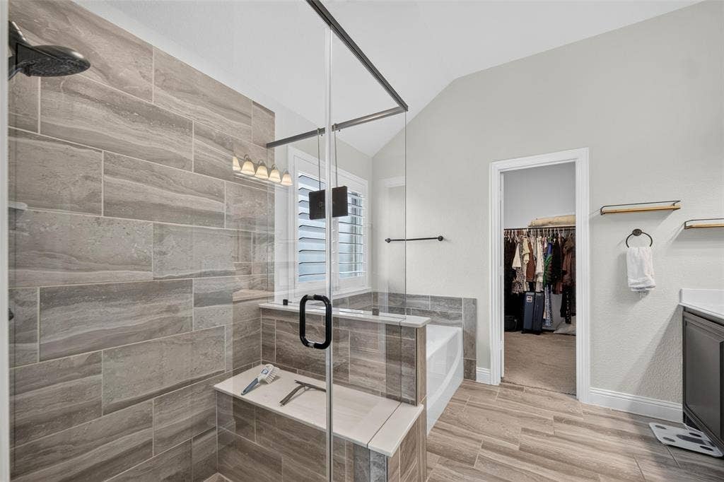 3210 Creekhaven Drive, Melissa, TX 75454 Walk-in shower featuring glass paneling, floor-to-ceiling stone-finish tile, and built-in bench seating, complemented by wood-finish floor tiling and a soaking tub