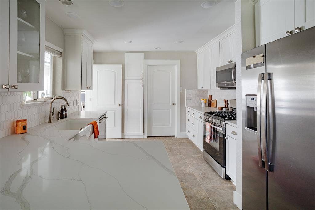 Kitchen featuring white cabinetry with brushed nickel hardware, stainless steel appliances, a white farmhouse sink, patterned tile backsplash, and light-toned countertops with veining