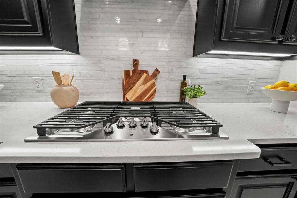 2628 Camden Court, Celina, TX 75009 Stainless steel gas cooktop with heavy-duty grates, light-toned stone-finish countertops, linear tile backsplash, and dark shaker-style cabinetry with under-cabinet lighting