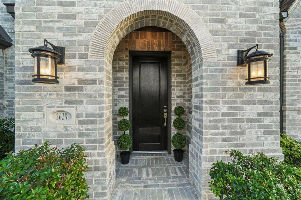 2628 Camden Court, Celina, TX 75009 Entryway featuring a grey brick facade, arched stone surround, solid dark-finish entry door, and two wall-mounted metal lanterns