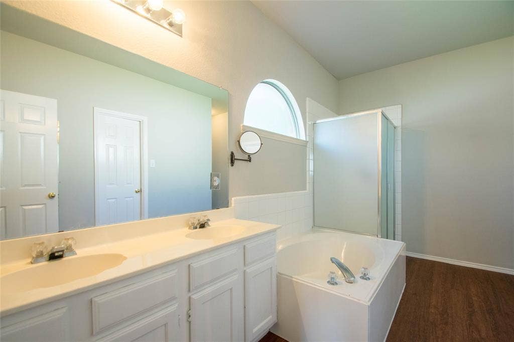 Dual vanity with integrated sinks, large mirror, wood-finish flooring, built-in soaking tub, and shower enclosure with frosted glass