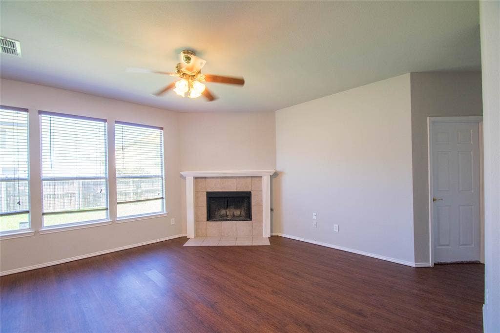 Spacious living area featuring wood-finish flooring, multiple windows with blinds, a ceiling fan, and a tiled fireplace with a white mantel
