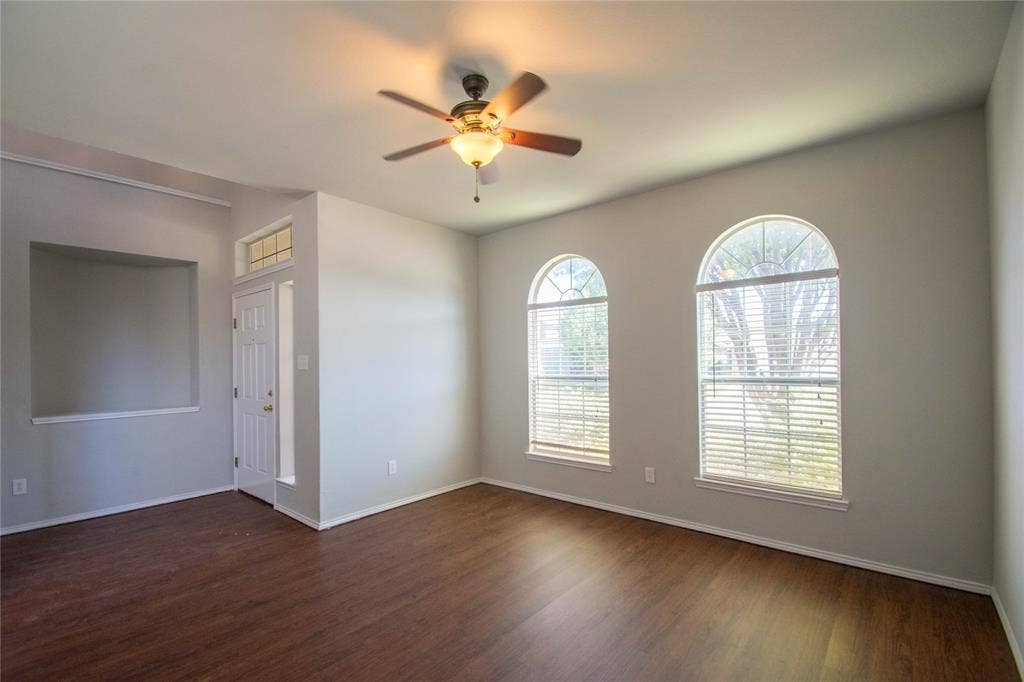 Wood-finish flooring, two arched windows with blinds, a ceiling fan with integrated lighting, a front entry door with transom window, and a wall niche