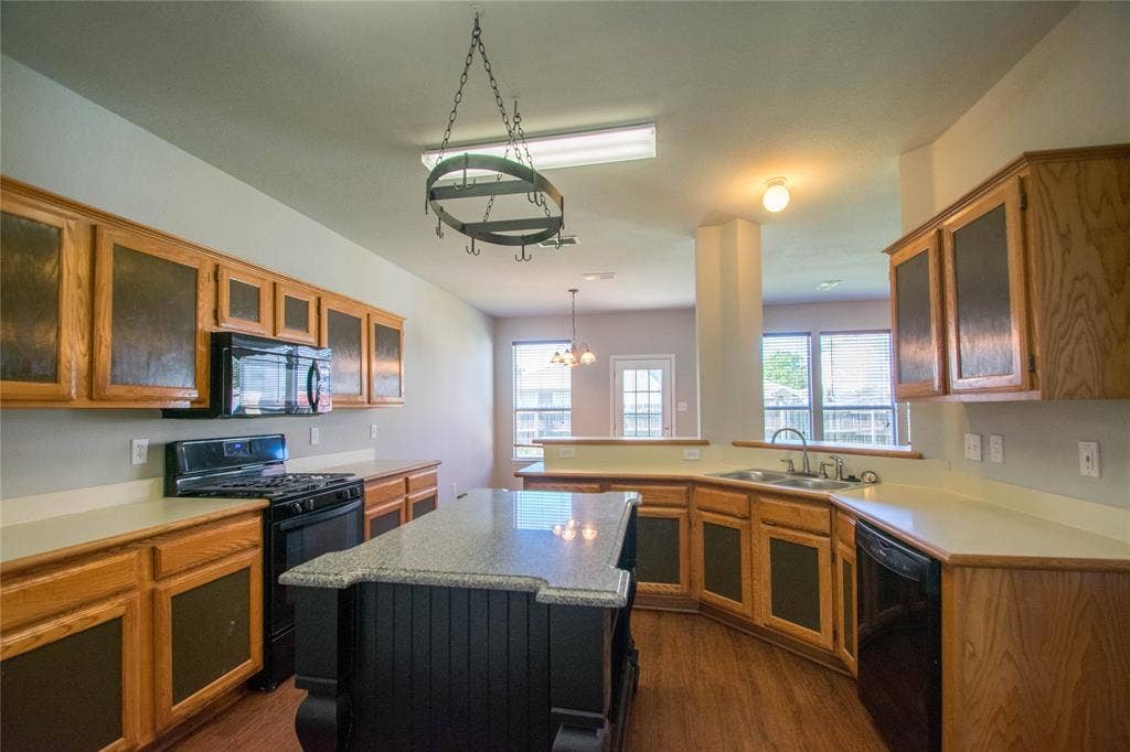 Spacious kitchen featuring wood cabinetry with glass-front uppers, stainless steel appliances, quartz countertops, a large center island with seating, hardwood flooring, and a decorative wrought-iron pendant fixture suspended from the ceiling