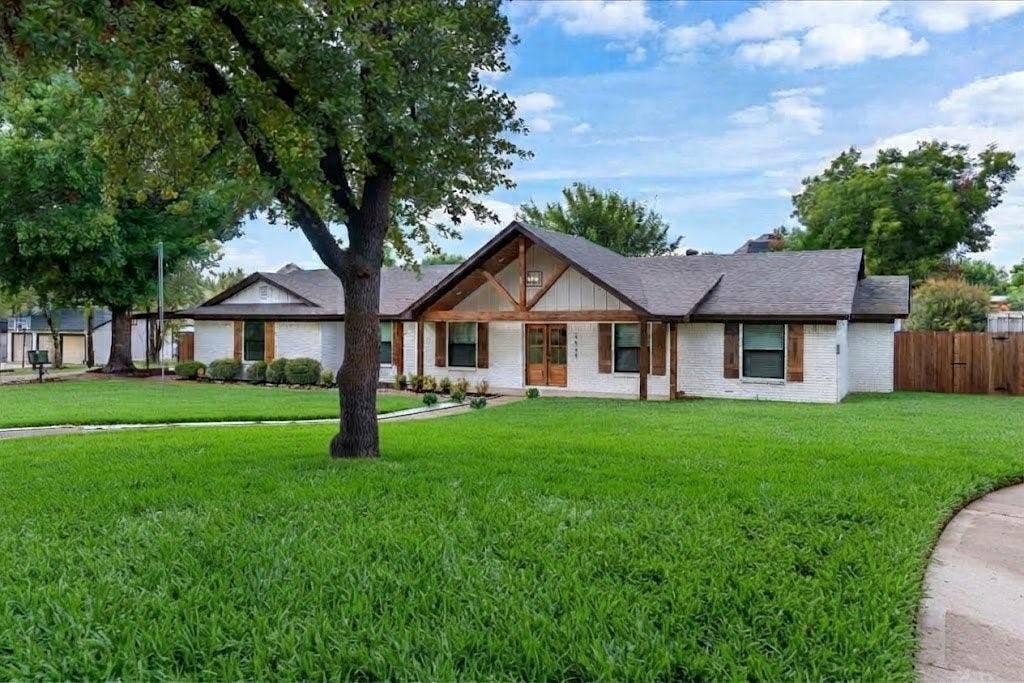 White exterior with dark roof shingles, front porch with wood beam accents, wood double entry doors, wood window shutters, and a front yard with established landscaping