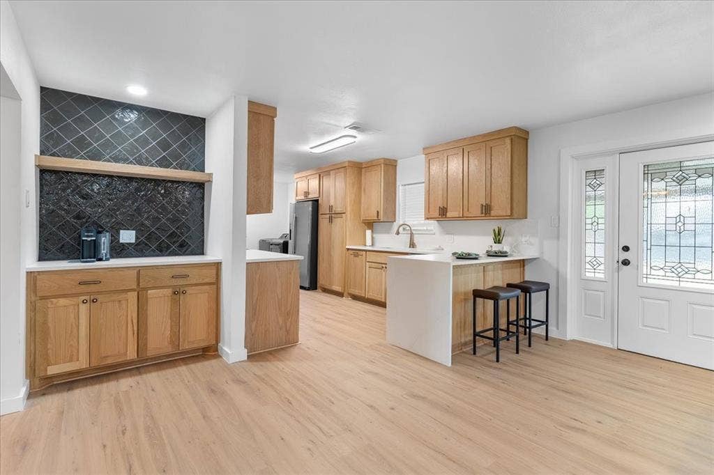 Wood-finish flooring, light wood cabinetry, white countertops, black tile backsplash, and a decorative glass front door