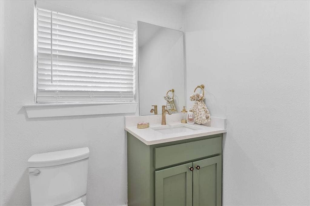 White wall-mounted toilet, vanity with sage green cabinetry, white countertop, integrated sink, and a rectangular mirror