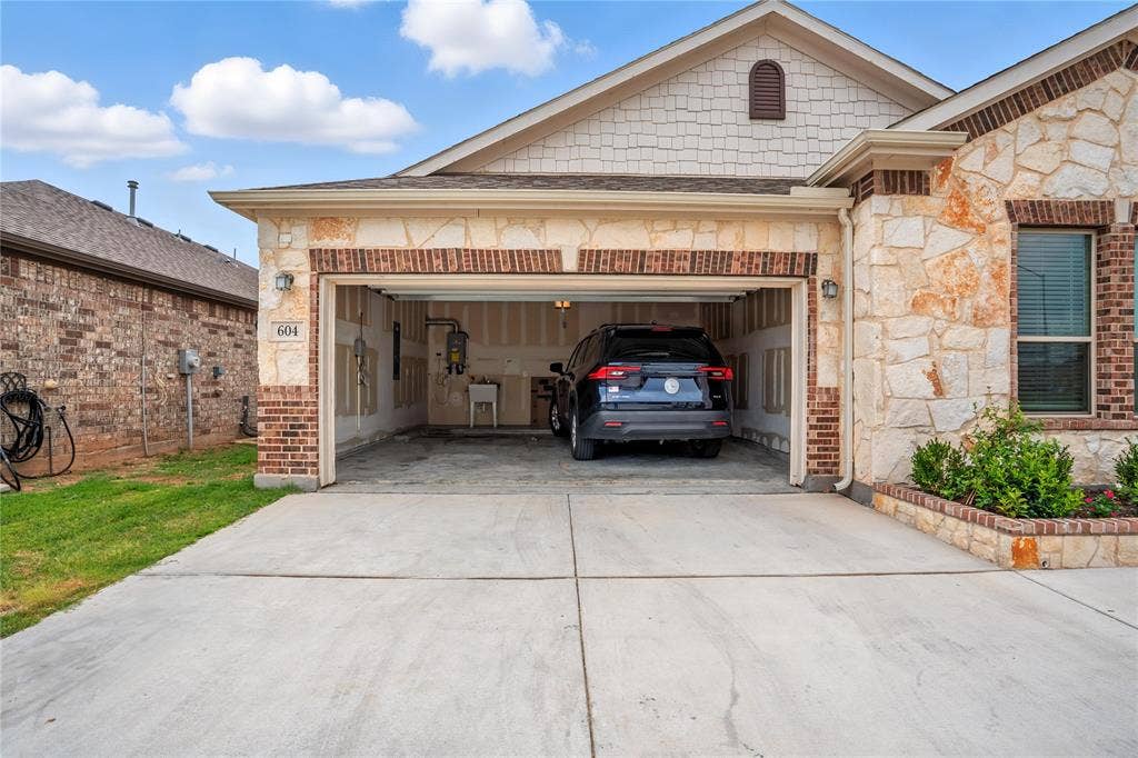 View of front facade with driveway, stone siding, brick siding, a garage, and roof with shingles