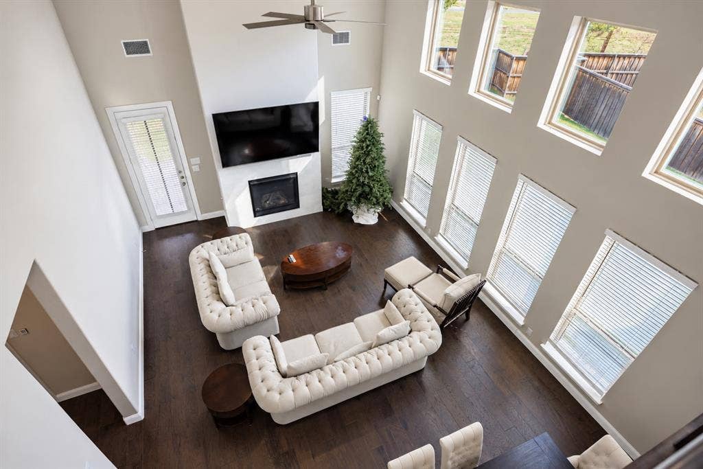 1603 Coventry Court, Farmers Branch, TX 75234 Soaring ceilings with a ceiling fan, multiple windows with blinds, a fireplace with a white surround, and dark wood-finish flooring