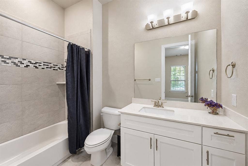 1603 Coventry Court, Farmers Branch, TX 75234 Bathroom featuring a white vanity with an integrated sink, a large wall-mounted mirror, a three-light vanity fixture, a toilet, and a tub with tile surround and decorative accent band
