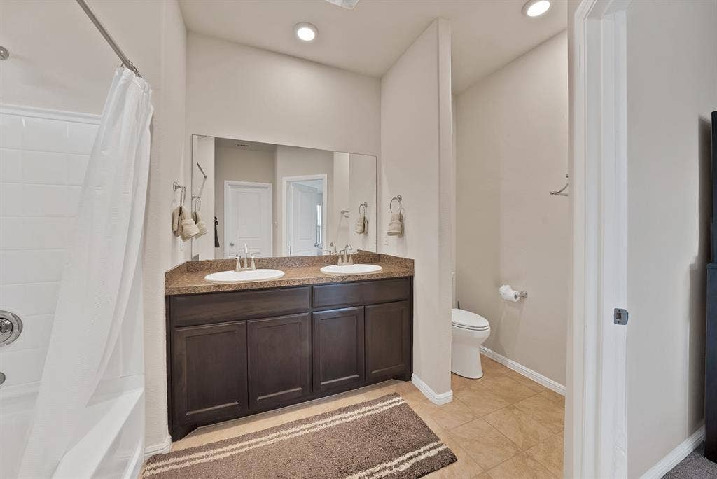 228 Foxhunter Street, Fort Worth, TX 76131 Dual vanity with integrated sinks, dark wood-finish cabinetry, a large wall-mounted mirror, white tub with shower, and tiled flooring